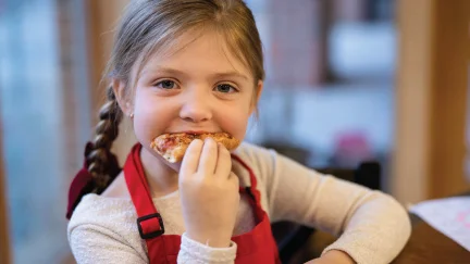 young girl eating slice of pizza smiling and wearing red RMHC Dayton apron